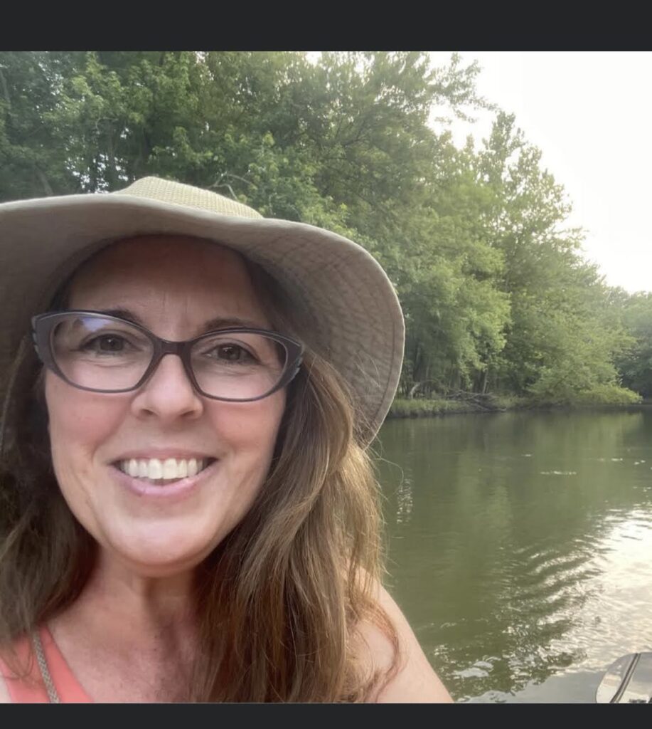 Terina Carter, a lady with long, dark hair, a hat, and glasses, smiiles as she poses in front of a river.